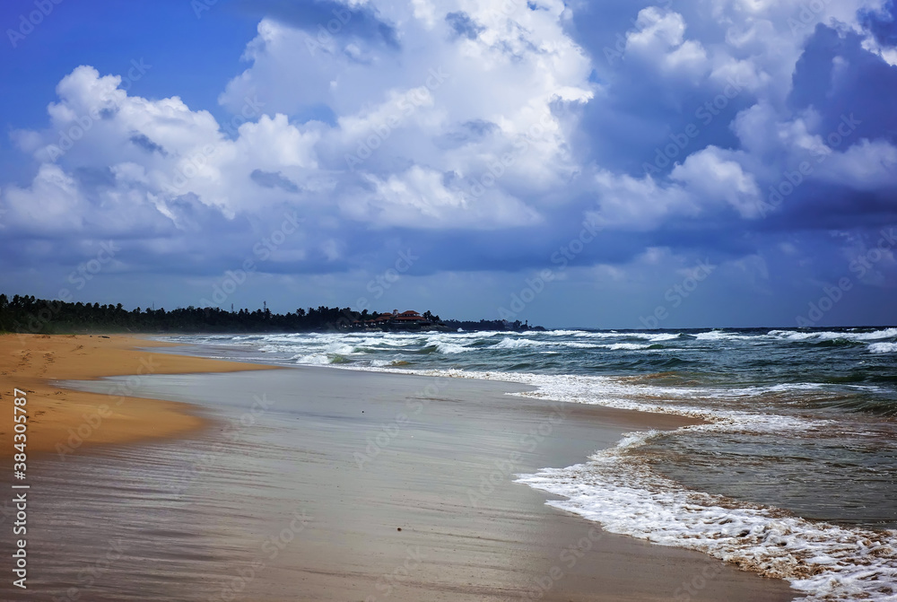 Foamy ocean waves roll onto the beach. Wet sand glistens. There are low picturesque clouds in the sky. Trees grow in the distance. Sri Lanka. Summer.