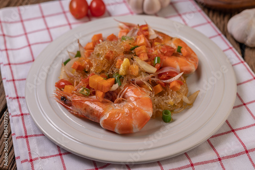 Stir-fried prawns with glass noodles in a white plate placed on a cloth with eggs and garlic.