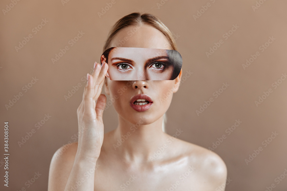 Shot of young woman with bare shoulders holding photo of her eyes with makeup in front of her face. Blonde girl opened her mouth sensuously Shot of young woman with bare shoulders holding photo of her eyes with makeup in front of her face. Blonde girl opened her mouth sensuously