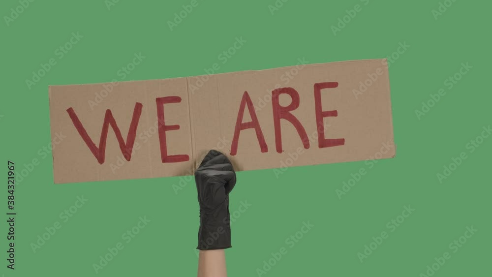 The hand of an unknown person in black gloves raise a cardboard poster WE ARE. Equality and unity concept. Isolated a green screen, chroma key. Close up. Slow motion.