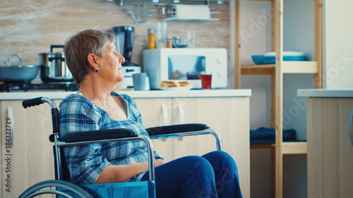 Handicapped senior woman in wheelchair standing alone in kitchen looking throug window. Pensive thoughtful lonely woman in solitude. Elderly disabled pensioner after injury and rehab, paralysis and