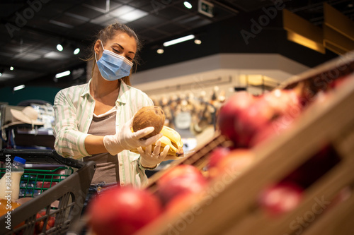 Woman with hygienic mask and rubber gloves and shopping cart in grocery buying fruit during covid-19 and preparing for a pandemic quarantine.