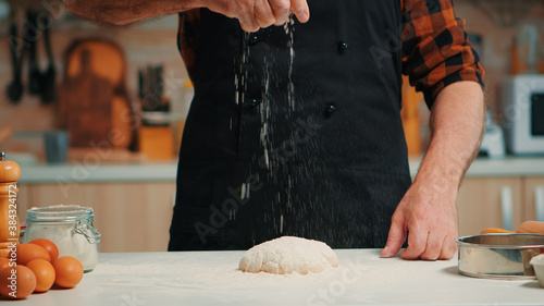 Senior bakery man sifting wheat flour on dough for kneading. Retired elderly chef with bonete and uniform sprinkling, sieving spreading rew ingredients with hand baking homemade pizza and bread. photo