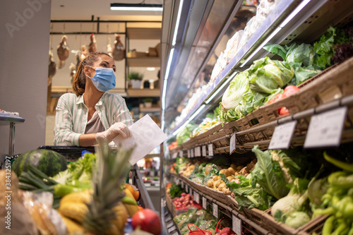 Woman with hygienic mask and rubber gloves and shopping cart in grocery buying vegetables during covid-19 and preparing for a pandemic quarantine.