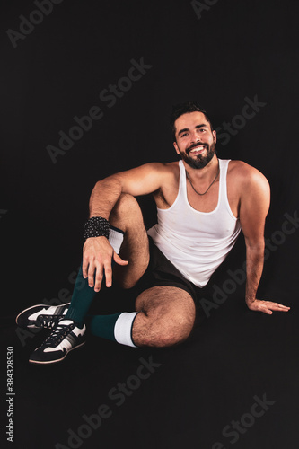 Young handsome break dancer man poses smiling in sportswear sitting on the floor on a black background