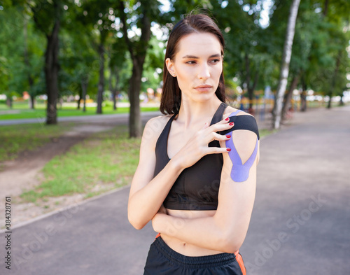 Young stunning muscular woman wearing black sports outfit. Fit brunette gorgeous female athlete putting colorful kinesiology tapes on shoulders. Concept of sports treatment, rehabilitation.