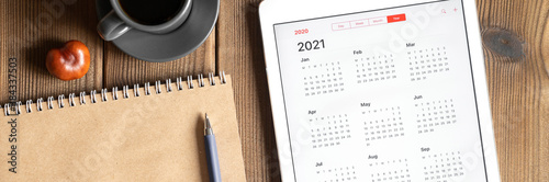 a tablet with an open calendar for 2021 year, a cup of coffee, chestnuts and a craft paper notebook on a wooden boards table background. banner