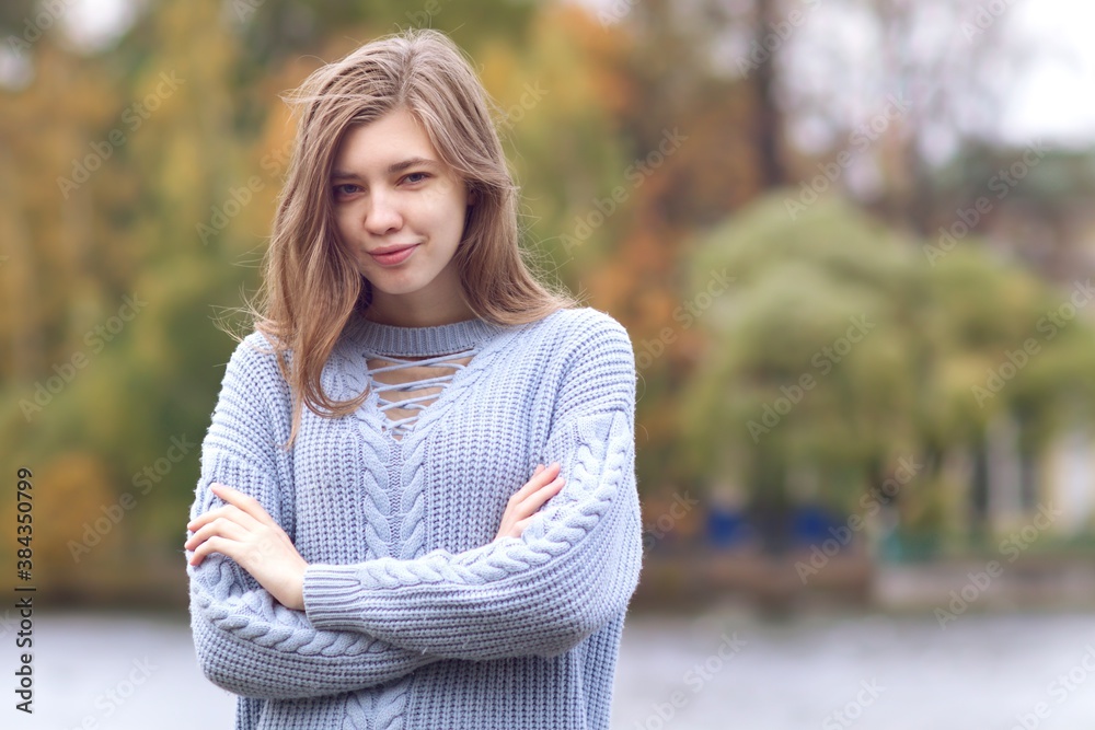 Obraz premium Portrait of young beautiful woman standing in autumn park in sweater with her hands crossed, smiling, looking at camera 