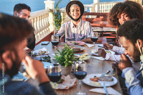 Happy multiracial friends eating and drinking wine together at dinner while wearing face masks during coronavirus pandemic outbreak - Social distance concept