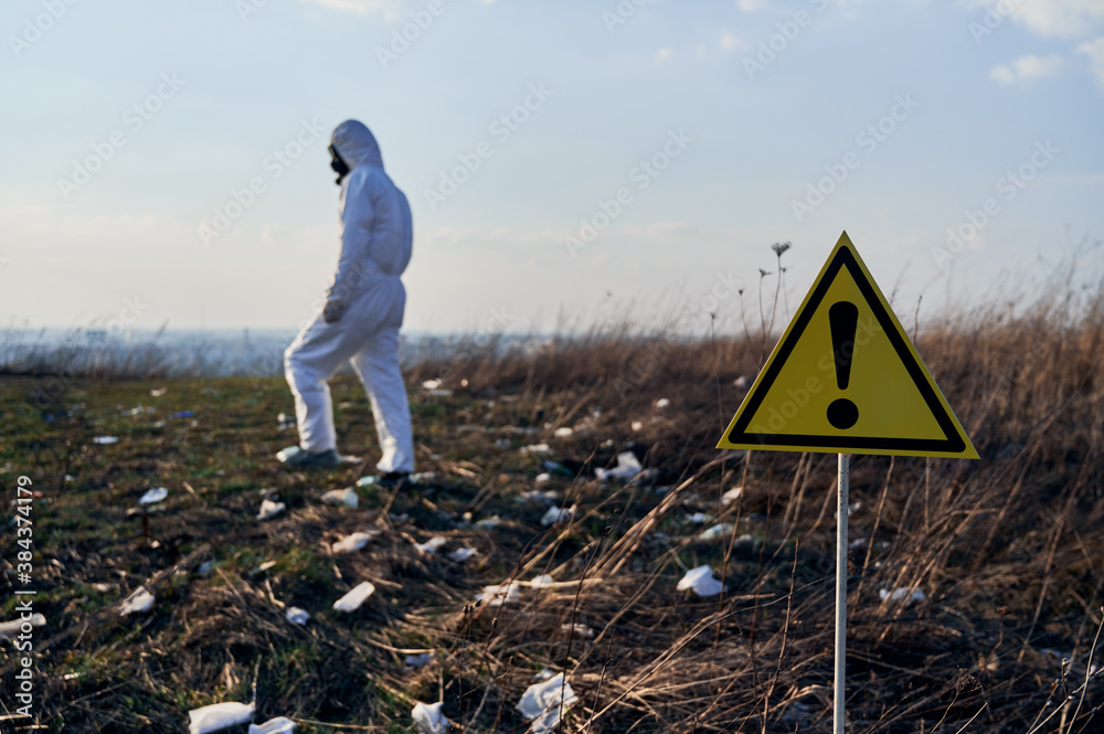 Obraz premium Focus on warning sign. Back view of male ecologist in protective suit walking in field with trash on blurred background. Concept of ecology, environmental pollution and danger.