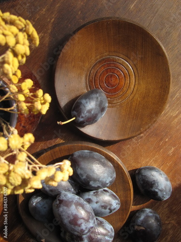 Autumn still life of plums on wooden plates and a wooden table, a vase with yellow dried flowers. Country style