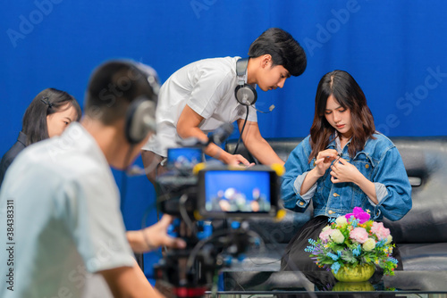 Television video camera recording interview in broadcast studio. Asian woman clipping tie-clip microphone and Professional cameraman with headphones looking video camera viewfinder. Selective focus