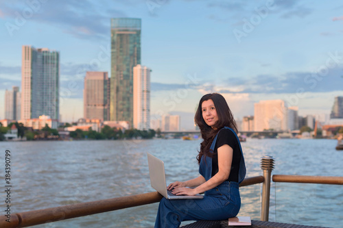 Asian woman holding laptop thinks looking for ideas or problems solutions in front of river beautiful view at sunset and background blur building skyscrapers.