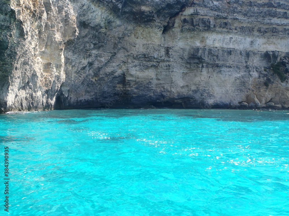 A photograph of a vibrant blue lagoon ocean with grey cliffs in Malta. Holiday feel, beautiful sunlight and clear water