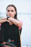 Young serious brunette female surfer with wristwatch standing behind surfboard