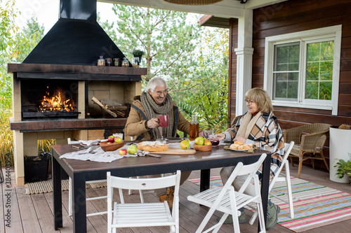 Cheerful senior couple in warm casualwear having tea by dinner table outdoors