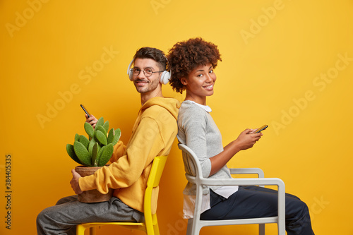 Fotografie Interracial friends pose on chairs against vivid yellow background hold mobile p