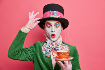 Surprised male hatter wears gentlemans clothes holds cup of hot tea pretends to come from wonderland wears vivid makeup celebrates halloween on costume party poses indoor alone. Body painting