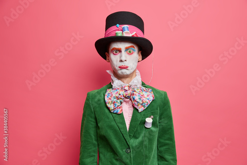 Discontent man dressed in halloween costume has professional makeup purses lips with offended expression poses indoor over rosy background. Halloween event or festival concept. Character of wonderland