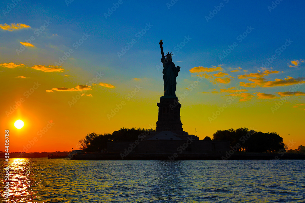 Statue of Liberty in silhouette at sunset viewed from the water with clouds and colorful sky.