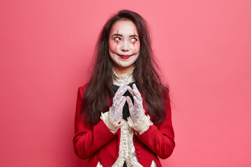 Scary brunette woman with zombie eyes and bloody scars dressed in halloween costume steepls fingers and has evil plan isolated over pink background. Horrible scary female on masquerade party