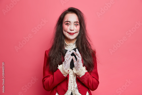 Happy vampire woman has devil plan and intention to do something wears halloween makeup and masquerade costume poses indoor against rosy background. Terrifying female has image of spooky zombie