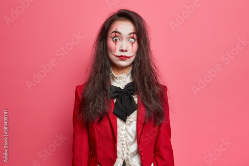 Shocked brunette woman with scars on face prepares for halloween festival stares bugged eyes tries to be dangerous isolated on pink background. Scarying horrible female vampire or zombie indoor
