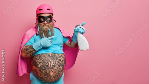 Superhero ready for spring cleaning. Plump bearded housekeeper in costume poses with detergent and indicates away on blank space over pink background. Domestic service. Professional clean up photo