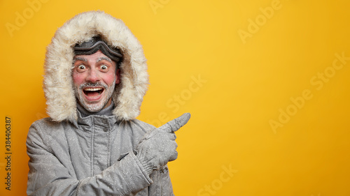 Happy frozen man in warm clothes has fun during winter points away on empty space wears gloves isolated on yellow background. Cheerful male adventurer with frosted beard and ski goggles has expedition