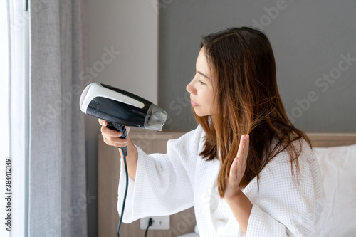 Young Asian girl in white bathrobe sitting on her bed and blow drying her hair in the morning