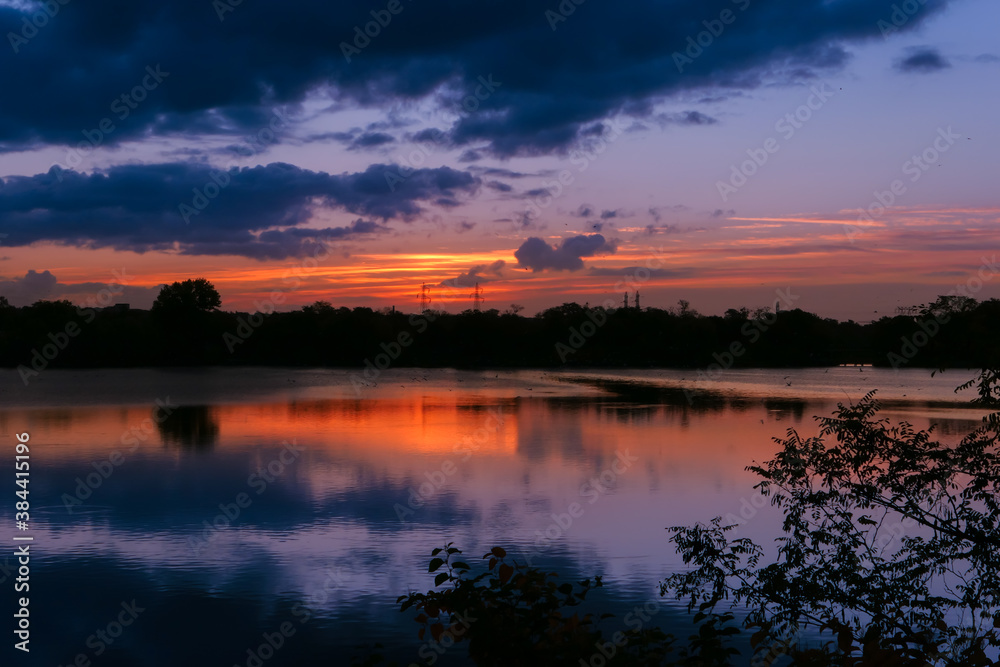 Amazing sunrise in rural scene. Symmetry of the sky in a lake at sunset. Clouds reflecting on the water. Quiet relaxing scene with a beautiful colorful cumulonimbus. Silhouette of vegetations.