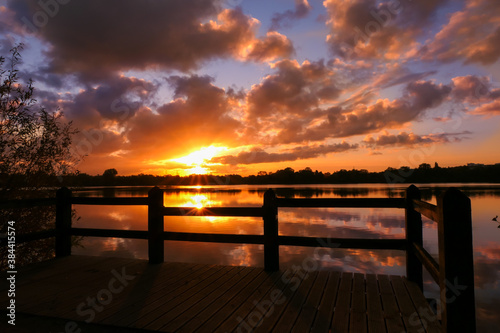 Amazing sunrise in rural scene. Symmetry of the sky in a lake at sunset. Clouds reflecting on the water. Quiet relaxing scene with a beautiful colorful cumulonimbus. Wooden pontoon in the foreground.