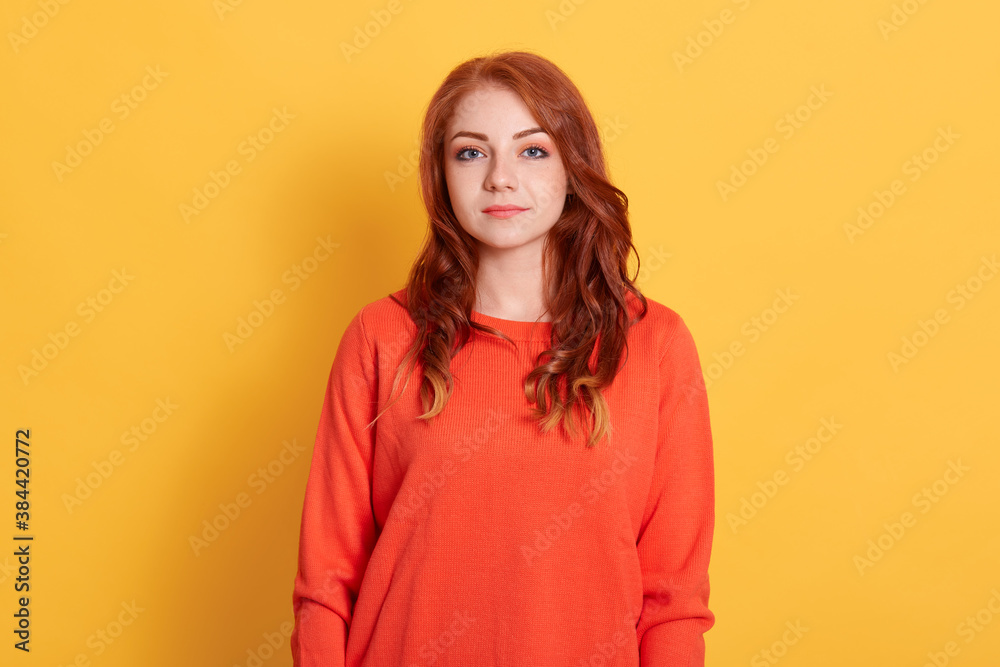 Portrait of Caucasian ginger woman with calm and serious face expression, looks directly at camera, wears orange jumper, has natural beauty, models against yellow wall. Portrait of Caucasian ginger woman with calm and serious face expression, looks directly at camera, wears orange jumper, has natural beauty, models against yellow wall.