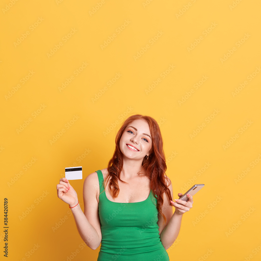 Happy beautiful lady with red hair and charming smile, being in good mood, having unlimited credit card, holding phone in hands, wearing green dress against yellow background. Copy space for promo. Happy beautiful lady with red hair and charming smile, being in good mood, having unlimited credit card, holding phone in hands, wearing green dress against yellow background. Copy space for promo.