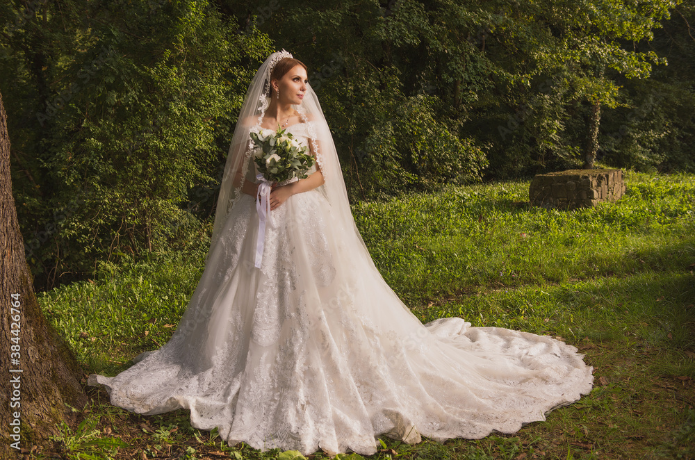 bride in a wedding dress with a long train and a bouquet against the background of ancient walls and nature