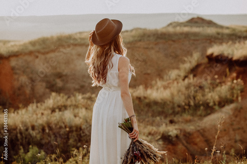 the bride walks on the field in the rays of the setting sun, the bride's white dress glows in the rays of the sun, the bride in a boho style hat, the concept of a wedding abroad