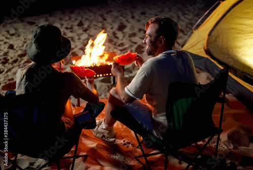 A cute woman and a handsome man are sitting on folding chairs near the tent by the fire, eating watermelon and having fun at night on the beach by the sea.
