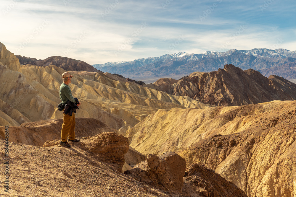 Hiker standing on the edge of a cliff high above a canyon of yellow rock with snow covered mountains in the distance, cloudy skies overhead, Golden Canyon, Death Valley National Park, California Hiker standing on the edge of a cliff high above a canyon of yellow rock with snow covered mountains in the distance, cloudy skies overhead, Golden Canyon, Death Valley National Park, California
