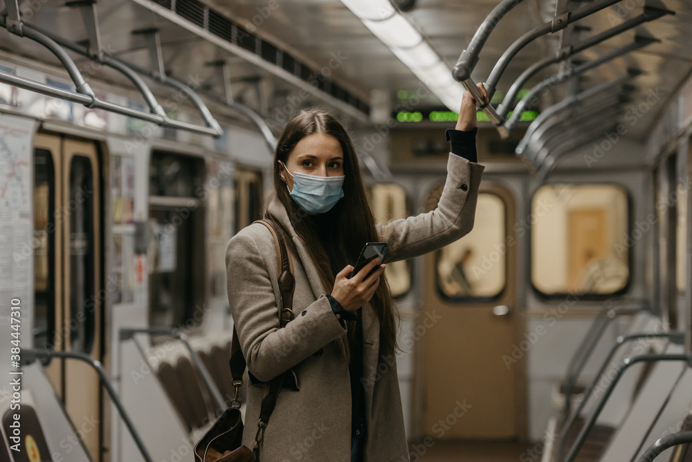 A woman in a medical face mask to avoid the spread of coronavirus is posing in a subway car. A girl with long hair in a surgical mask on her face against COVID-19 is holding a cellphone on a train.