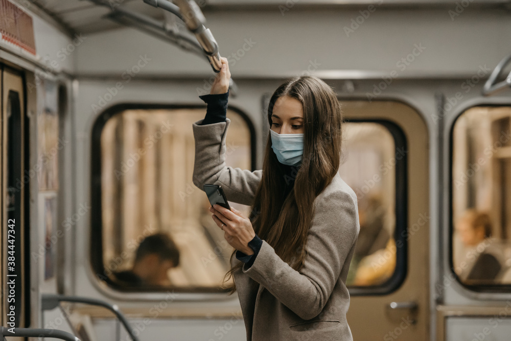 A woman in a face mask to avoid the spread of coronavirus is using a phone in a subway car. A girl with long hair in a surgical mask on her face against COVID-19 is holding a cellphone on a train.