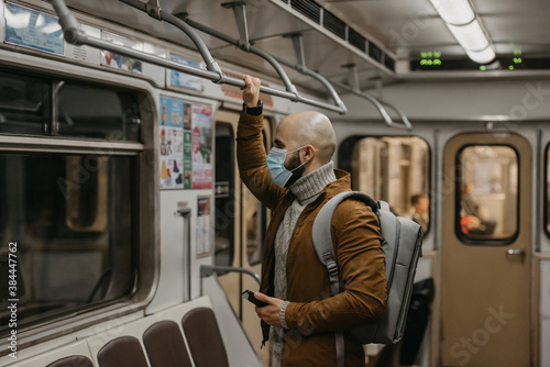 A man with a beard in a face mask to avoid the spread of coronavirus is looking through the window in a subway car. A bald guy in a surgical mask against COVID-19 is holding a phone on a metro train.