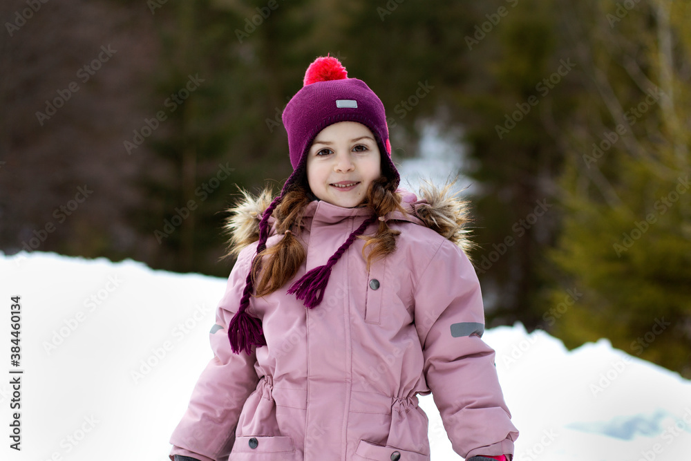 Cute little girl on the background of a forest of winter fir trees. Ski resort. Child in overalls. Winter walks, fun outdoors winter time.