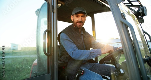 Billede på lærred Portrait of young Caucasian male farmer in cap sitting in tractor with open door and smiling to camera