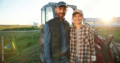 Portrait Caucasian happy couple of married farmers standing in hugs at tractor, looking at each other and smiling to camera. Outdoor. Man hugging woman in field. Agriculture concept. Wife and husband.