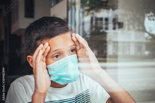 Young ill boy looking sad with protective mask at home behind window in quarantine and lockdown missing school and freedom during Covid-19 Coronavirus worldwide pandemic