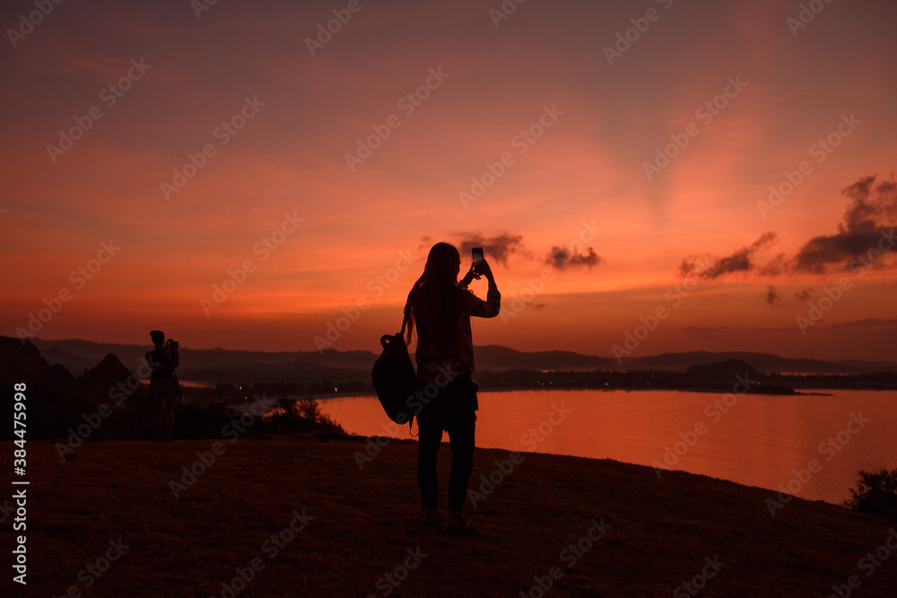 Silhouette woman take picture sunset by the sea on mobile phone. Tourist with backpack photographing ocean red sunset sky