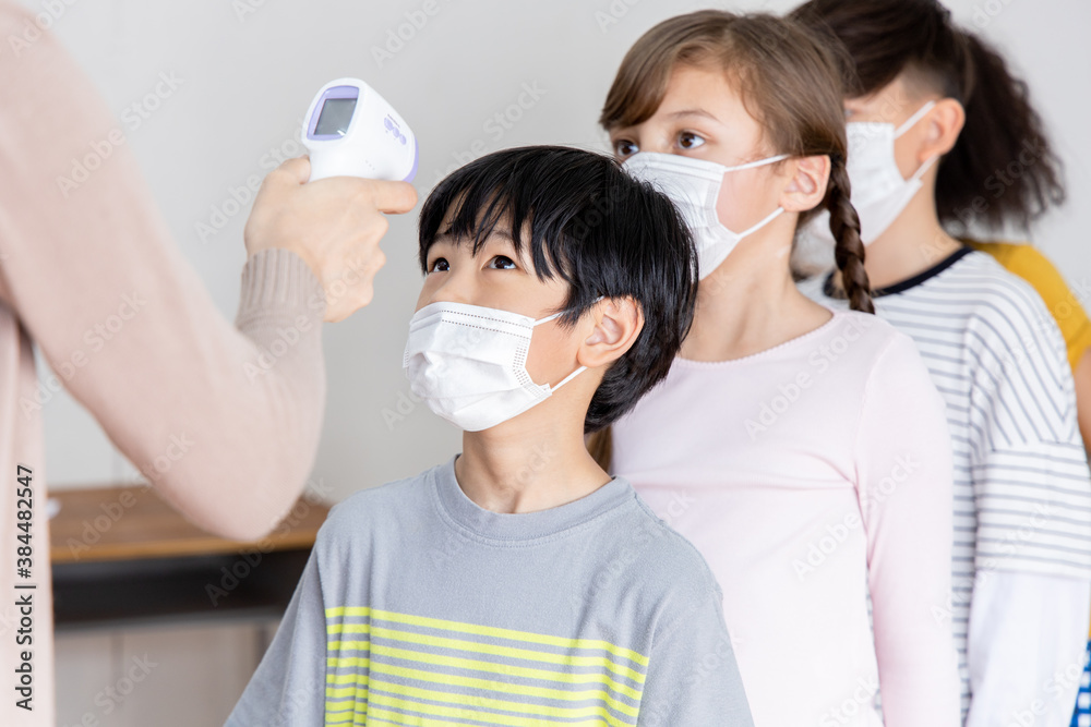 A group of Kids students wearing masks lined up waiting for woman teachers to Check Fever by Digital Thermometer in the classroom for Scan and Protect from Coronavirus Outbreak - Healthcare Concept