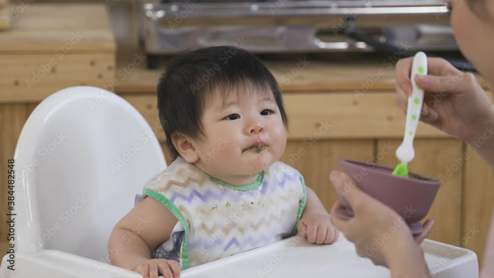 over the shoulder shot lovely toddler on the baby chair at home is enjoying her solid food from spoon served by her mom at lunchtime. over the shoulder shot lovely toddler on the baby chair at home is enjoying her solid food from spoon served by her mom at lunchtime.