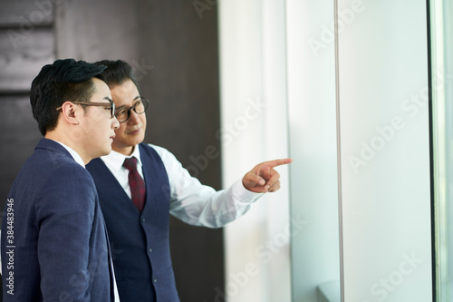 two asian business men standing by office window having a discussion