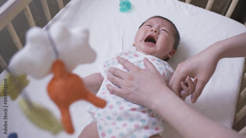 chinese mother is soothing her restless and crying newborn child by gently touching her on the chest and head in the crib with a colorful mobile. chinese mother is soothing her restless and crying newborn child by gently touching her on the chest and head in the crib with a colorful mobile.